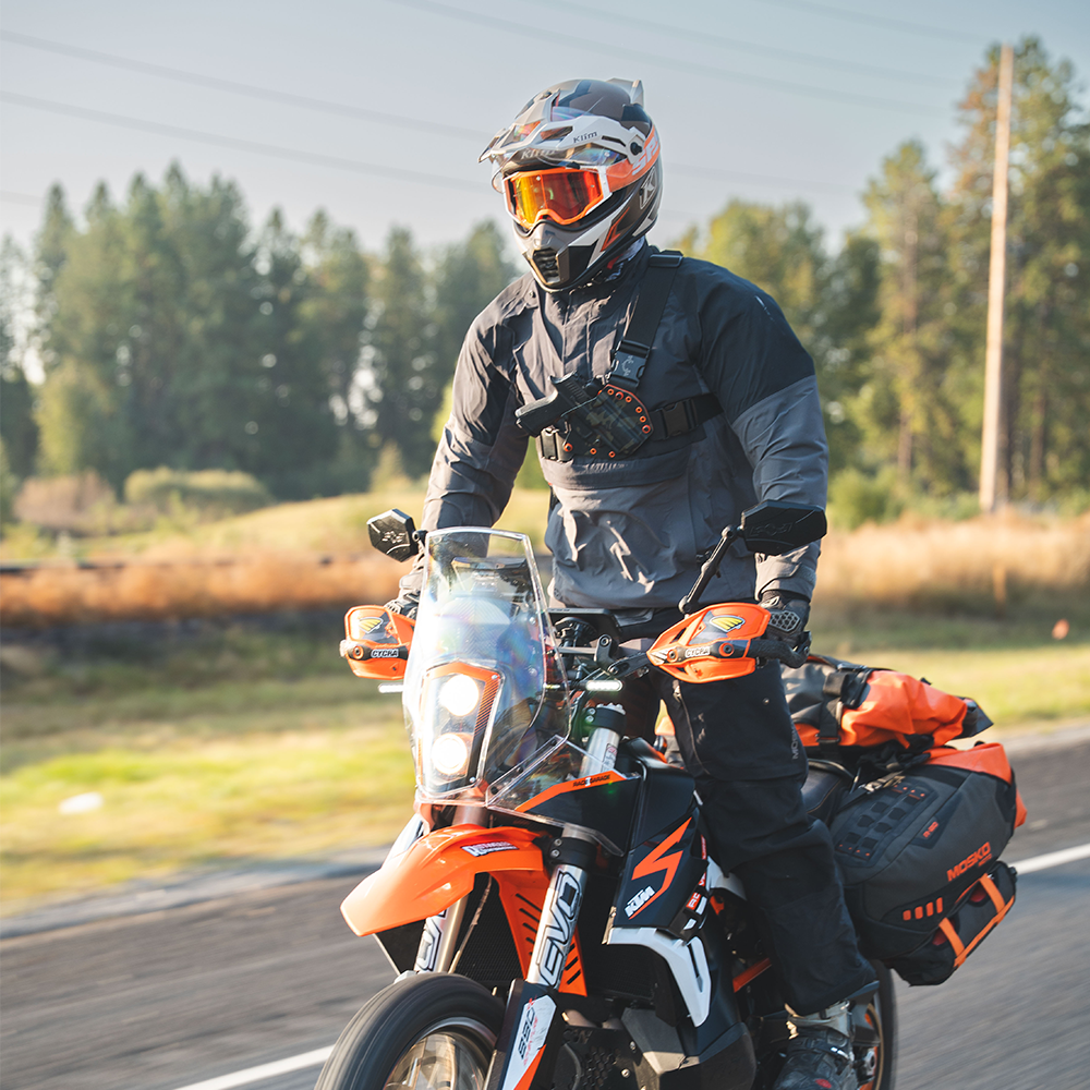 Person on an orange motorcycle with a helmet and gear, surrounded by trees. wearing a recon chest holster