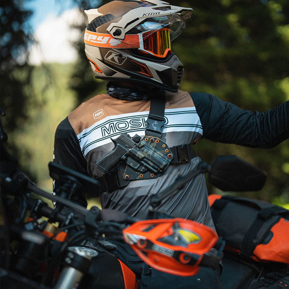 Person wearing a helmet and riding gear on a motorcycle with a forest background, wearing a Recon chest holster