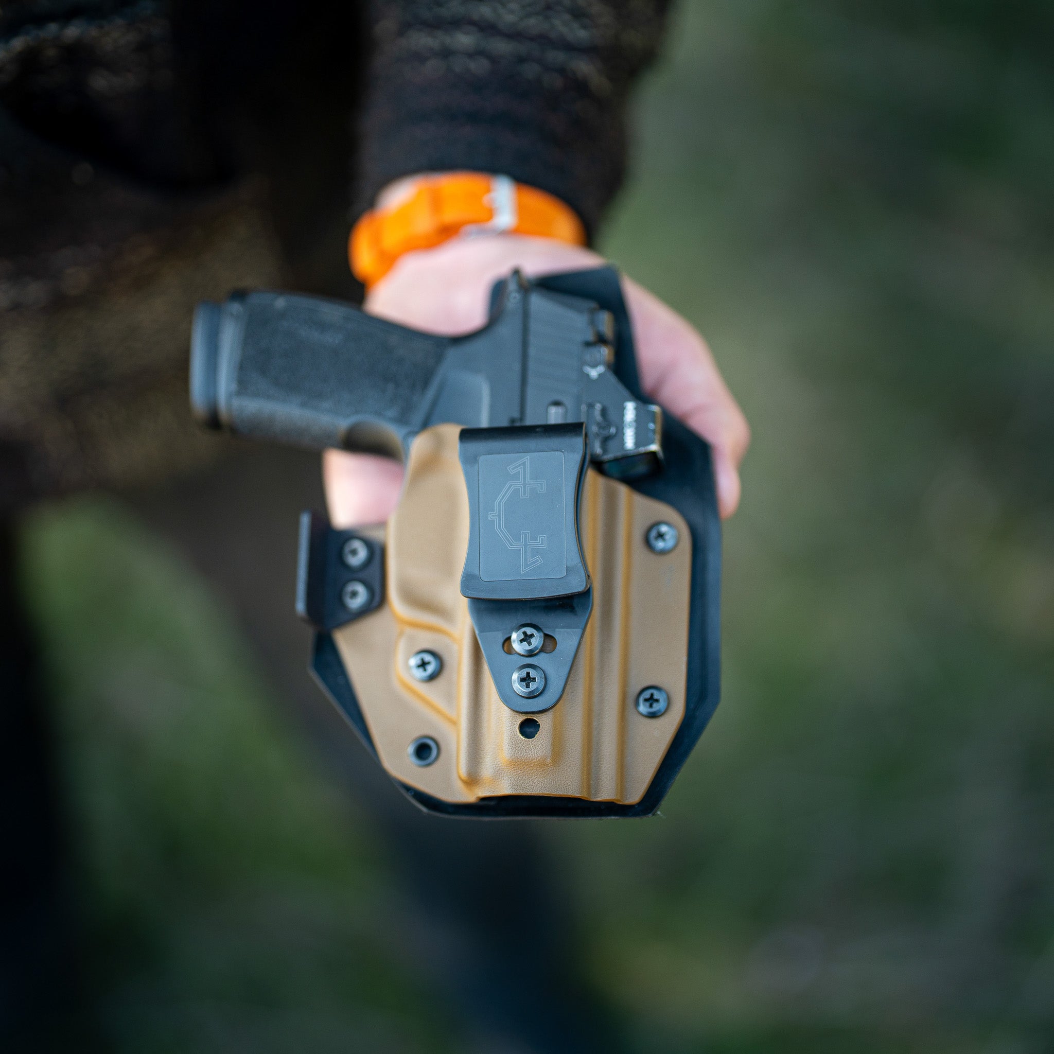 Hand holding a black and tan gun outlaw iwb(inside the waistband) holster with a blurred natural background