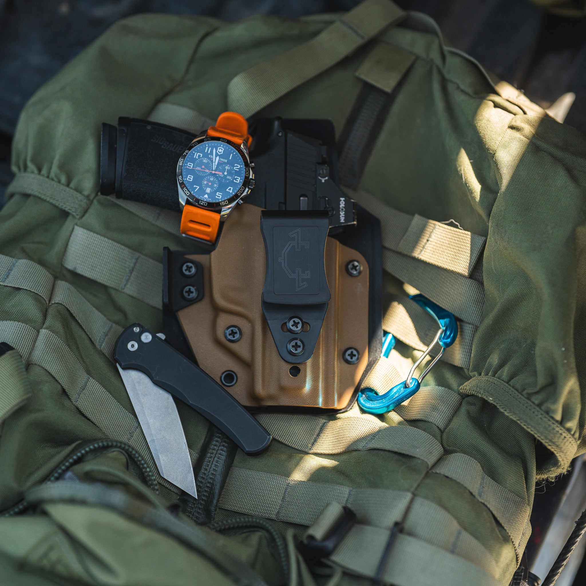 Green tactical bag with a knife, watch, and coyote colored outlaw IWB(inside the waistband) on a dark background