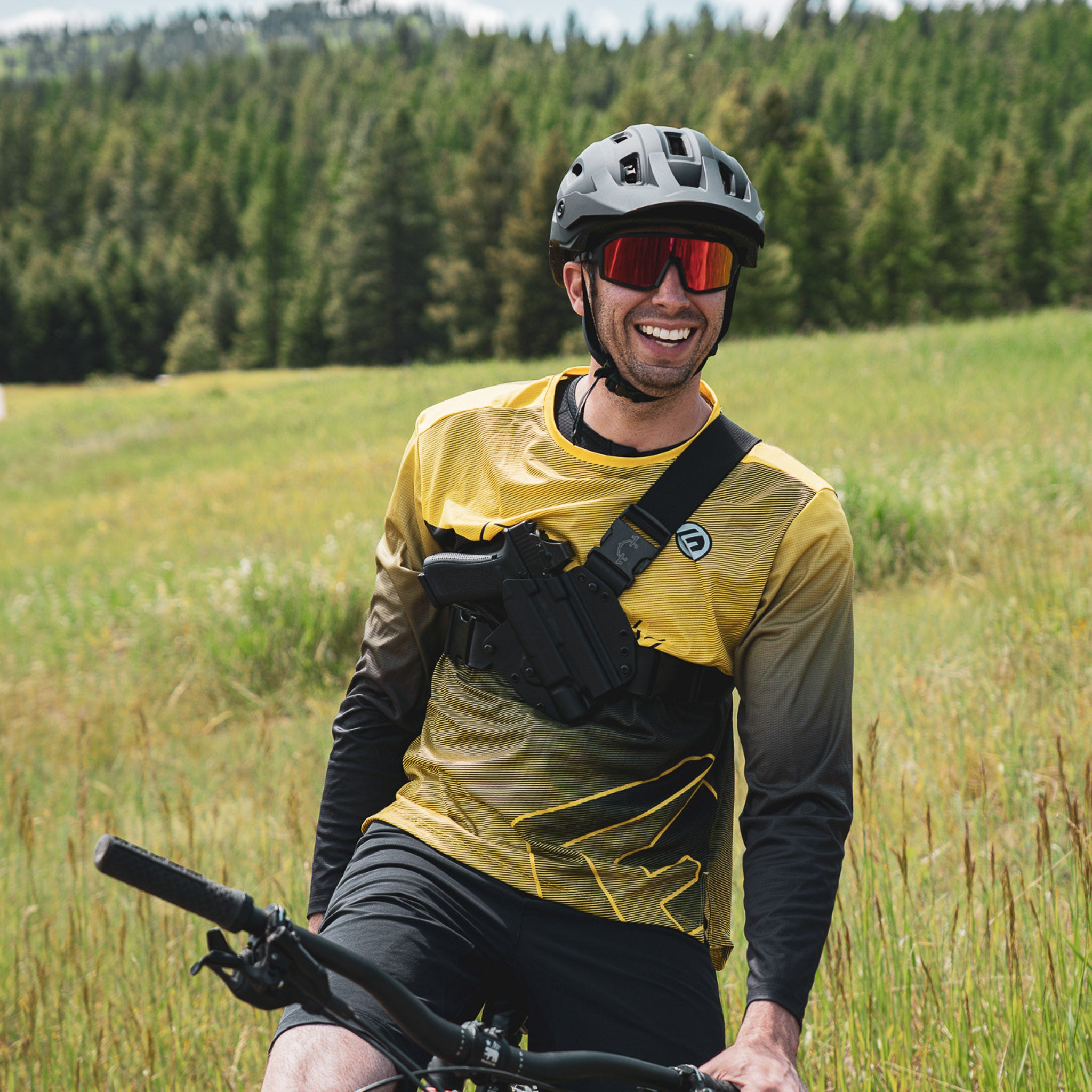 Man with a bicycle in a field with trees in the background with a recon chest holster with light options