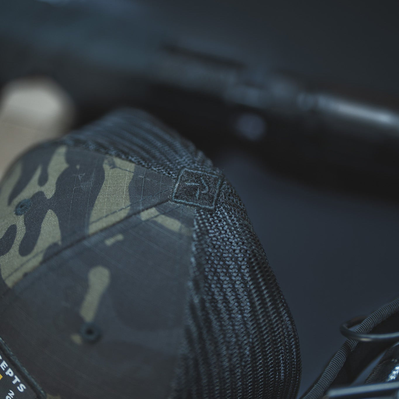 A close-up shot of a black camouflage trucker hat with a black mesh back, resting on a dark surface next to a firearm.