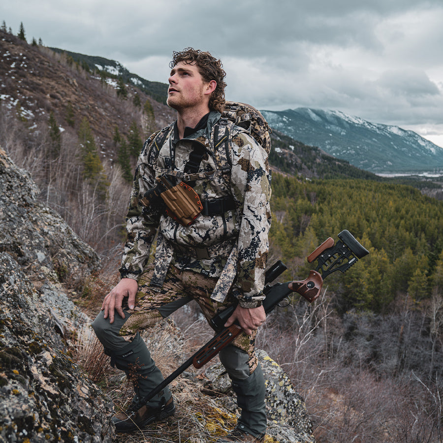Man in camouflage standing on a rock with a rifle, mountains in the background and wearing a recon chest holster