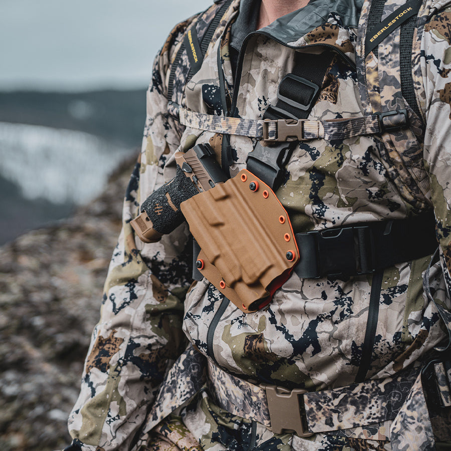 Person in camouflage gear with a Recon chest holster in coyote, on a rocky landscape.