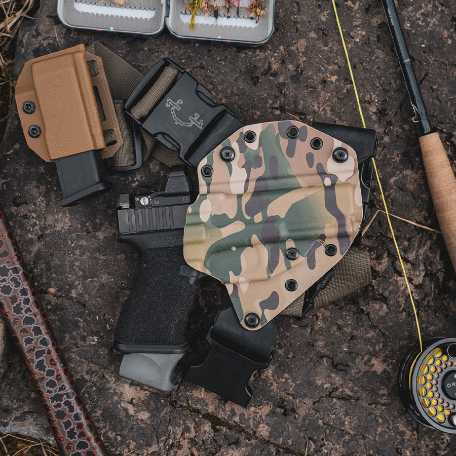 Camouflage gun recon chest holster on a textured surface with fishing equipment.