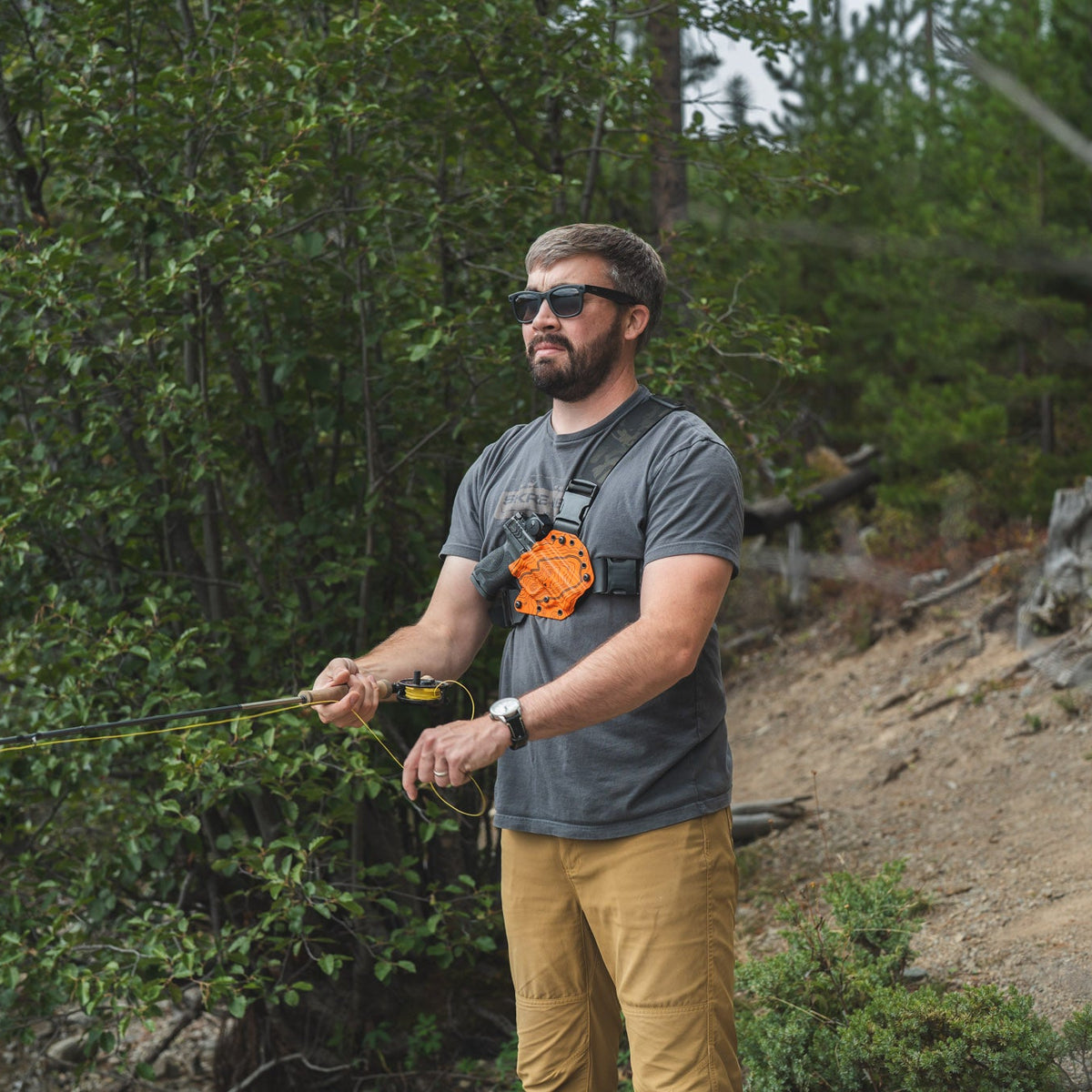 A man wearing a gray t-shirt, sunglasses, and tan pants stands in a wooded outdoor setting, holding a fishing rod with a chest-mounted orange device attached to his shirt.