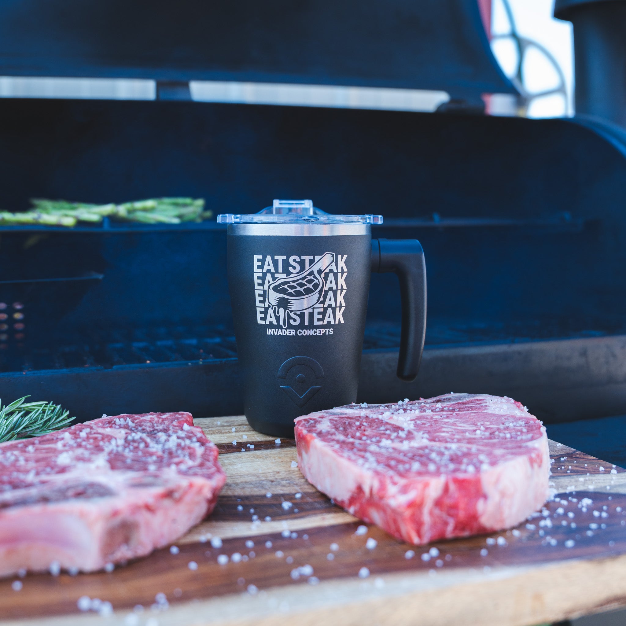 A black insulated mug with a clear lid stands on a wooden cutting board in front of a grill, with two raw steaks and coarse salt in the foreground. The mug is printed with the text 'EAT STEAK EVERY DAY' and a steak graphic, plus 'BRAZER CONCEPTS' near the bottom.