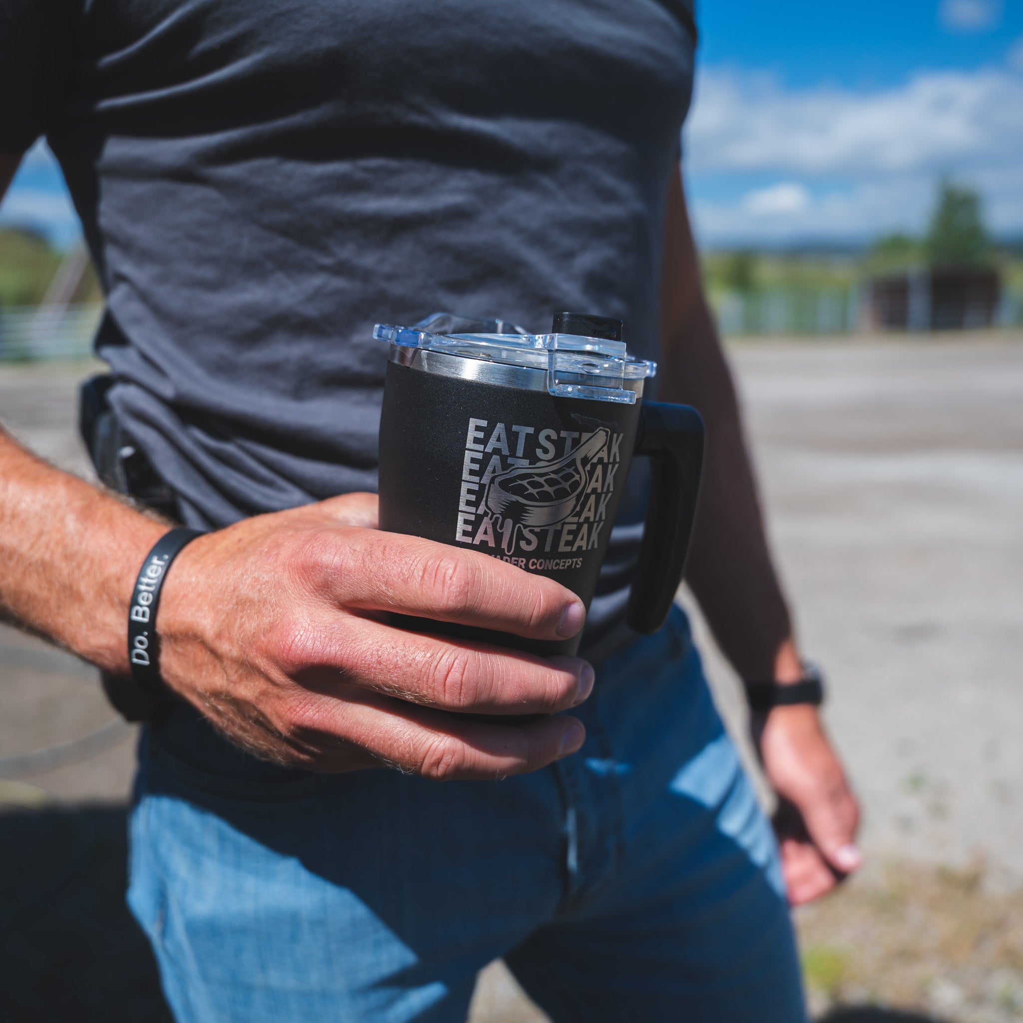 A person outdoors holding a black insulated tumbler with a clear lid. The tumbler has a white graphic that reads 'EAT STANLEY TEAM' with a bicycle illustration.