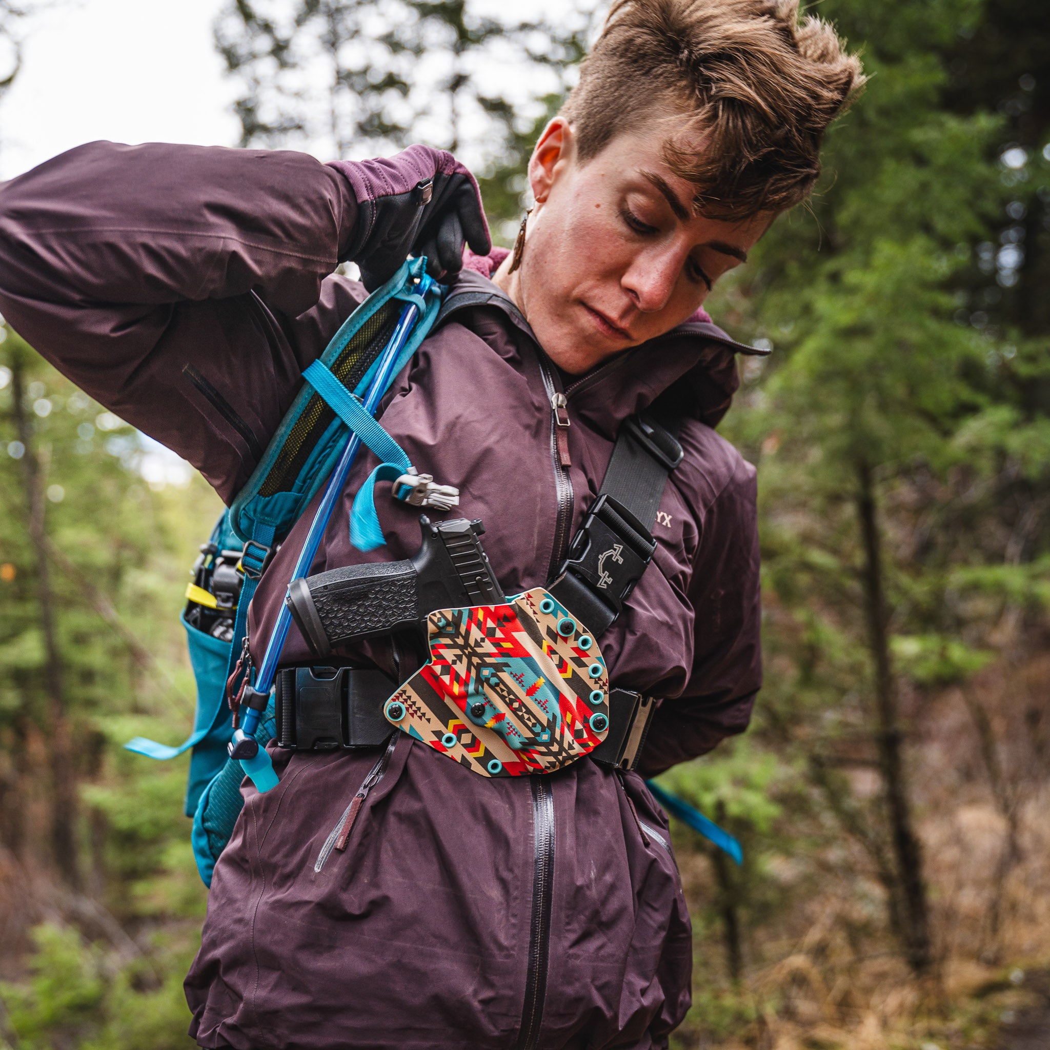 Woman in a purple jacket with a colorful backpack in a forest setting wearing a ladies recon chest holster