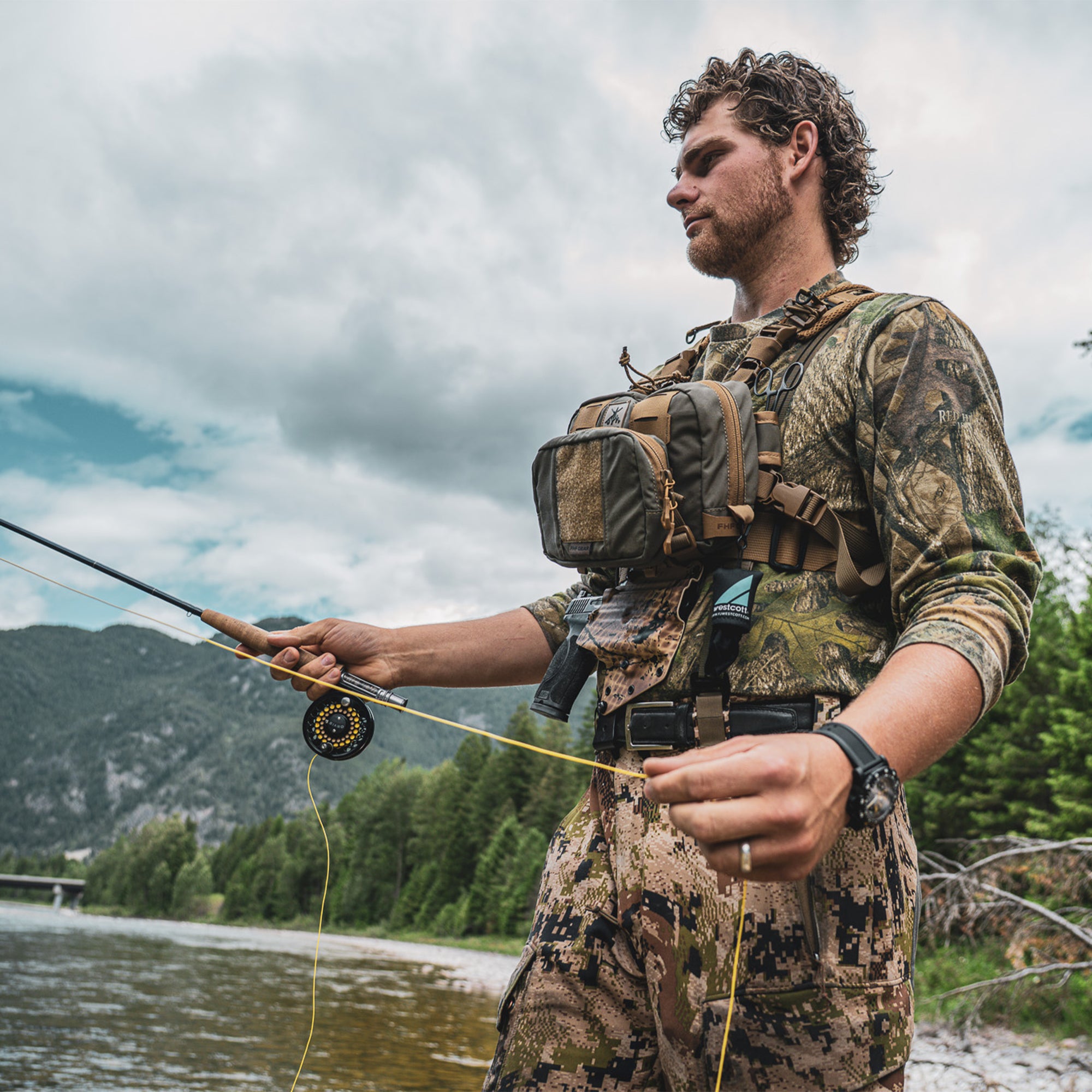 Man in camouflage clothing and gear holding a fishing rod by a river with mountains in the background while wearing a guide torso holster