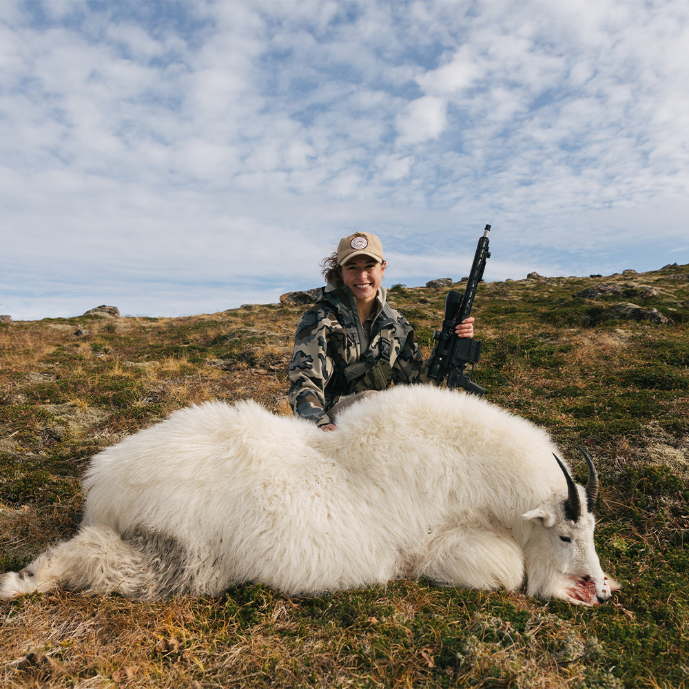 Woman holding a rifle next to a dead mountain goat on a grassy field with a cloudy sky and wearing a ladies recon chest holster