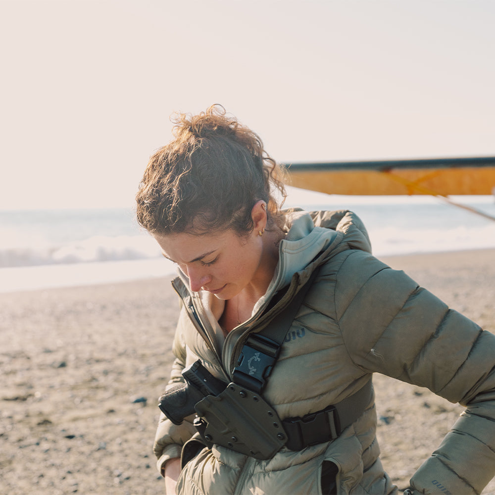 Lady wearing a green jacket with a ladies recon chest holster and harness on a beach