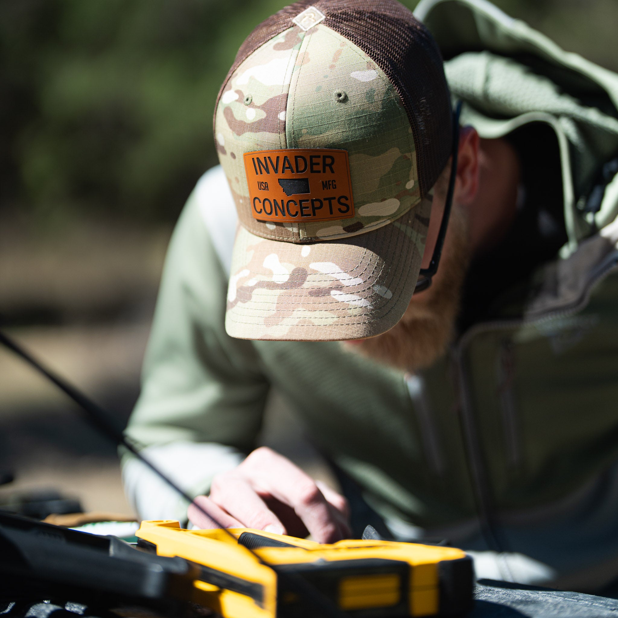 Person wearing a camouflage cap with 'Invader Concepts' logo, working on a yellow tool outdoors.