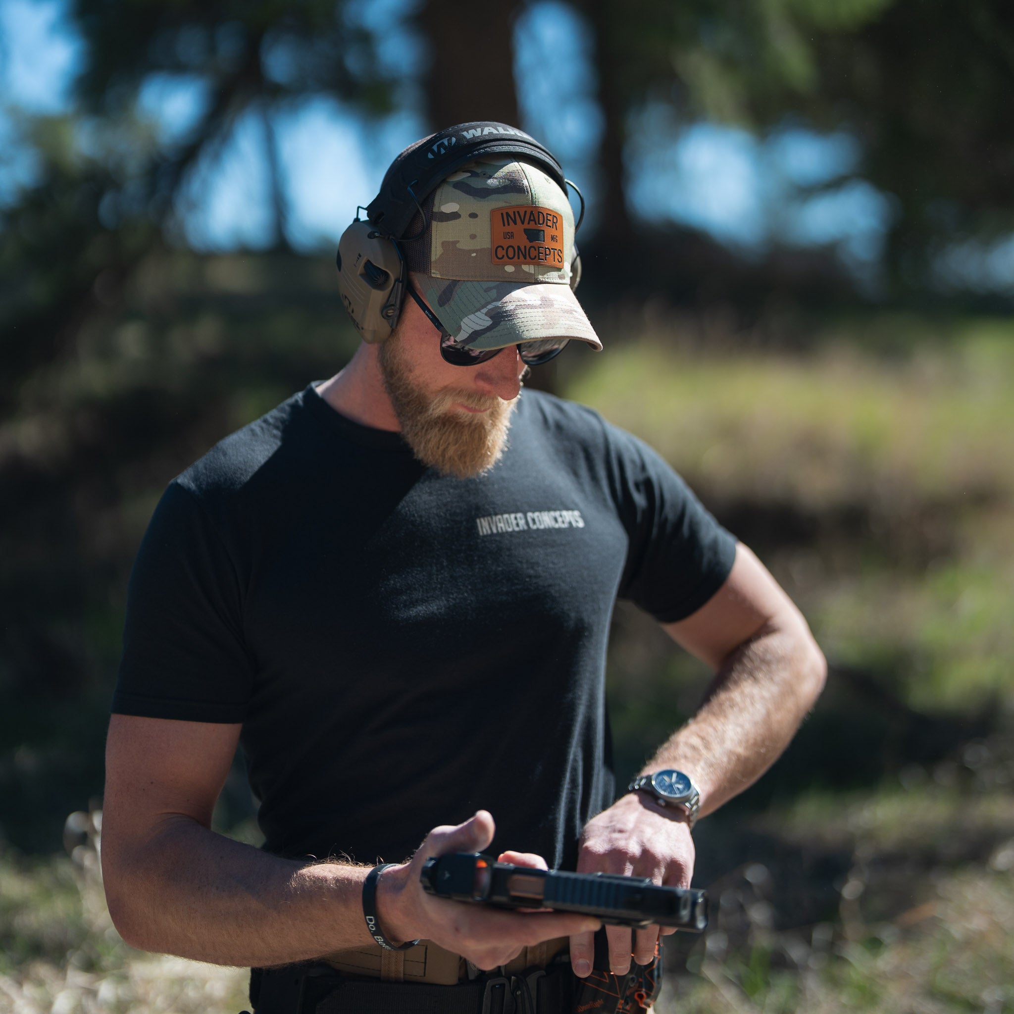 Man wearing a black t-shirt and camouflage cap with ear protection, holding a pistol in an outdoor setting.