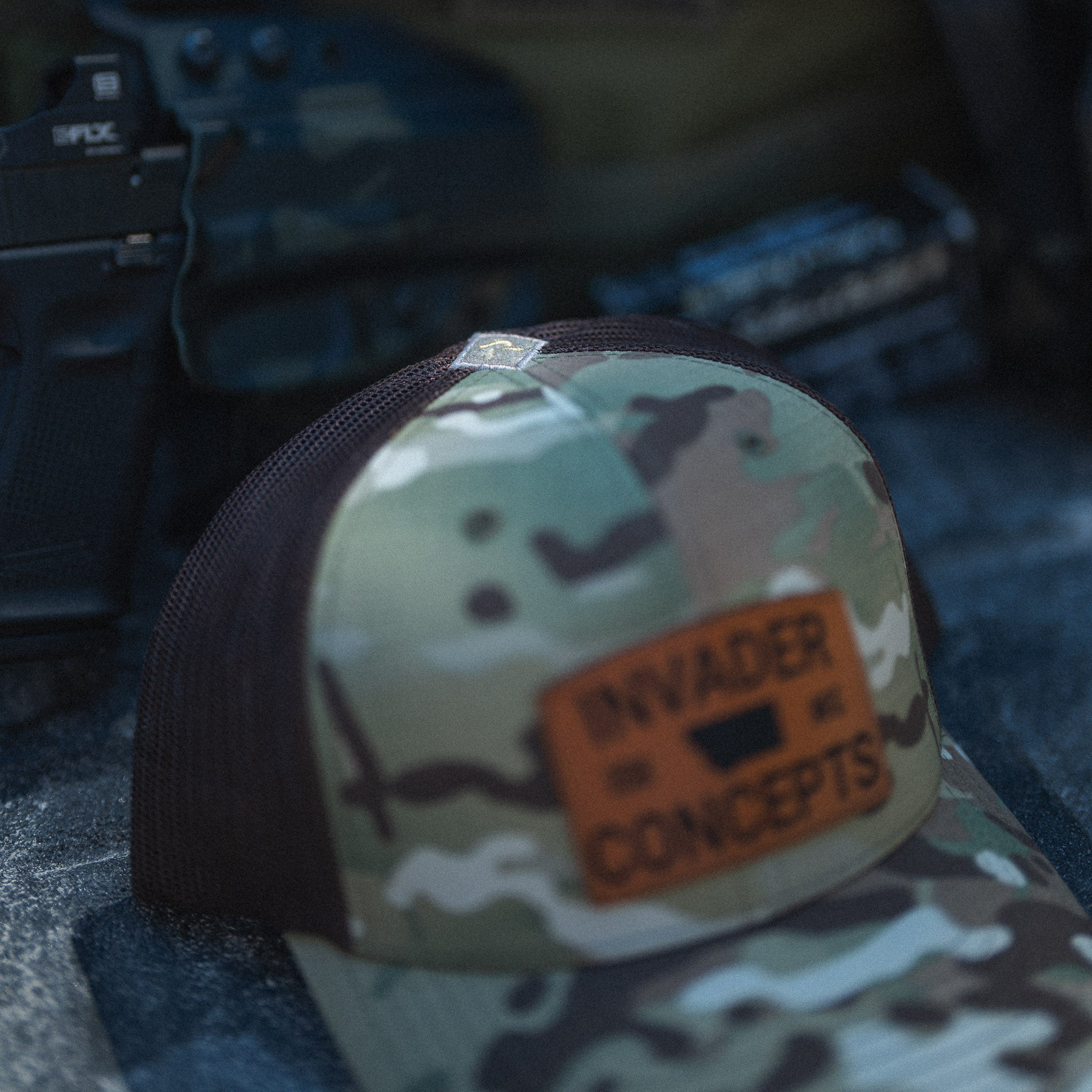 Camouflage cap with 'Invader Concepts' logo on a blurred background