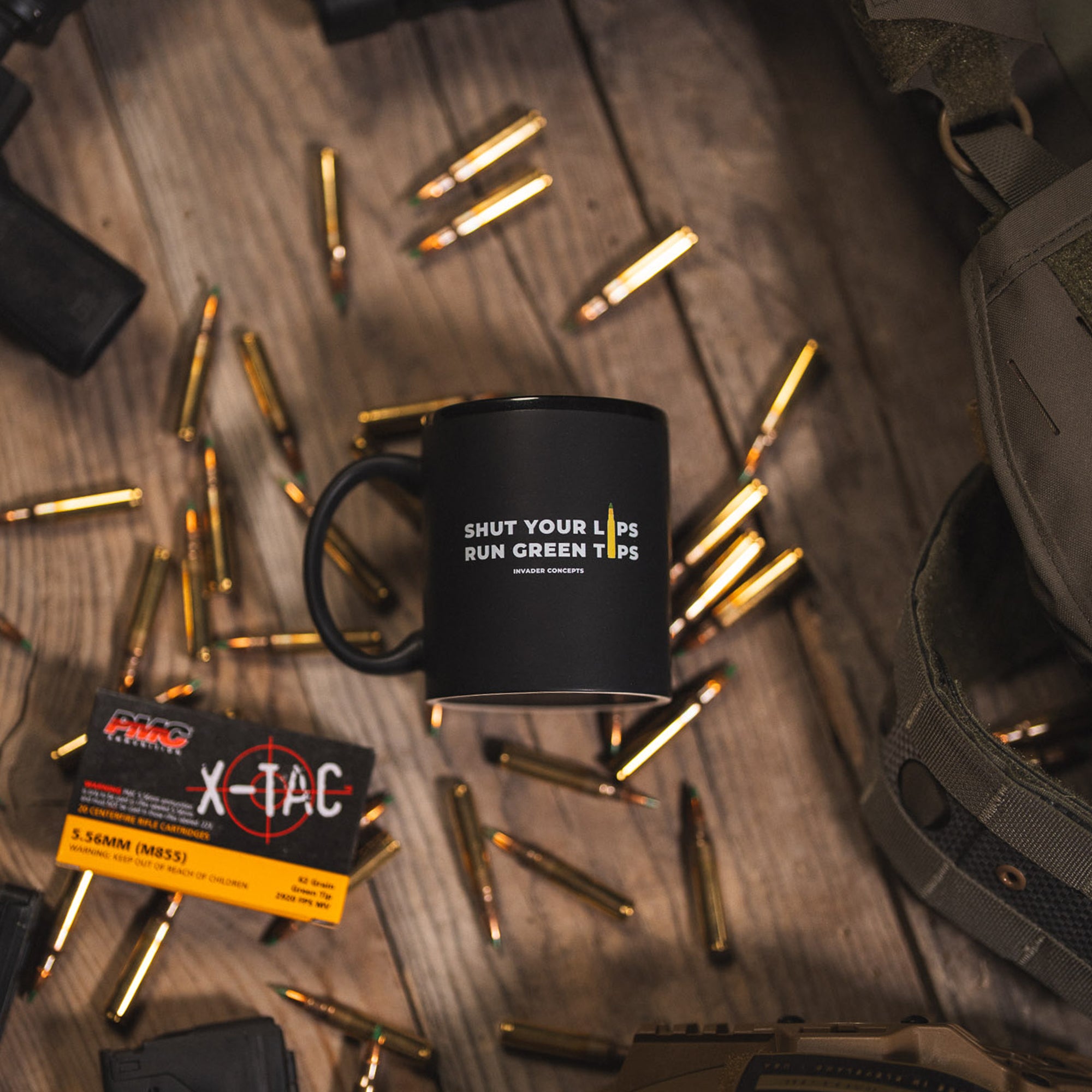 Black mug with text on a wooden surface with ammunition and tactical gear.