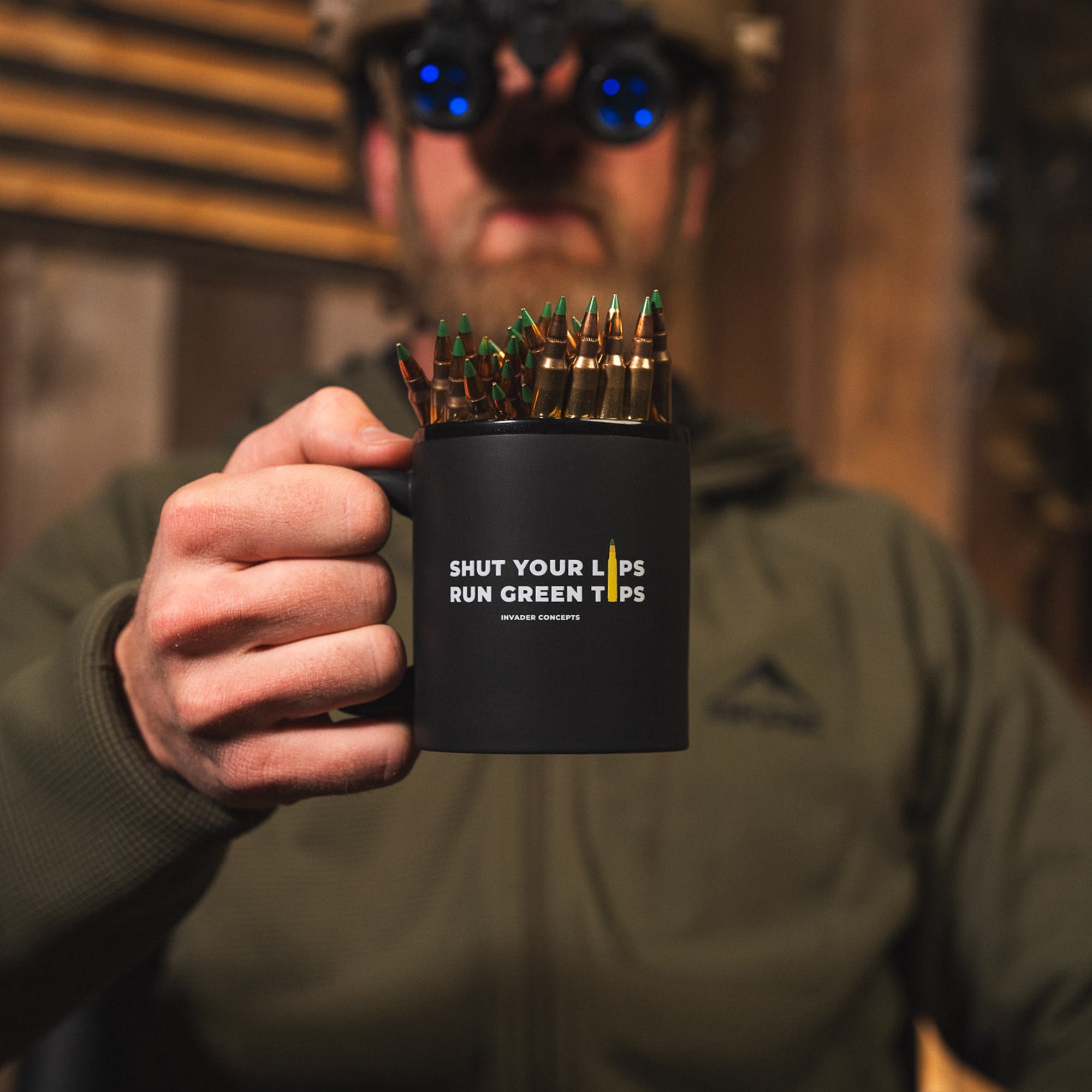 Person holding a mug with bullets and 'Shut Your Lips Run Green Tips' text, wearing goggles.