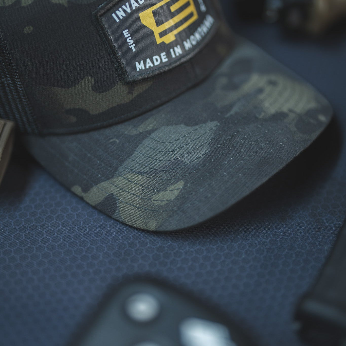 A close-up of a dark camouflage trucker cap with a rectangular patch reading 'INVADER CONCEPT' and 'MADE IN MONTANA' on a dark textured surface.