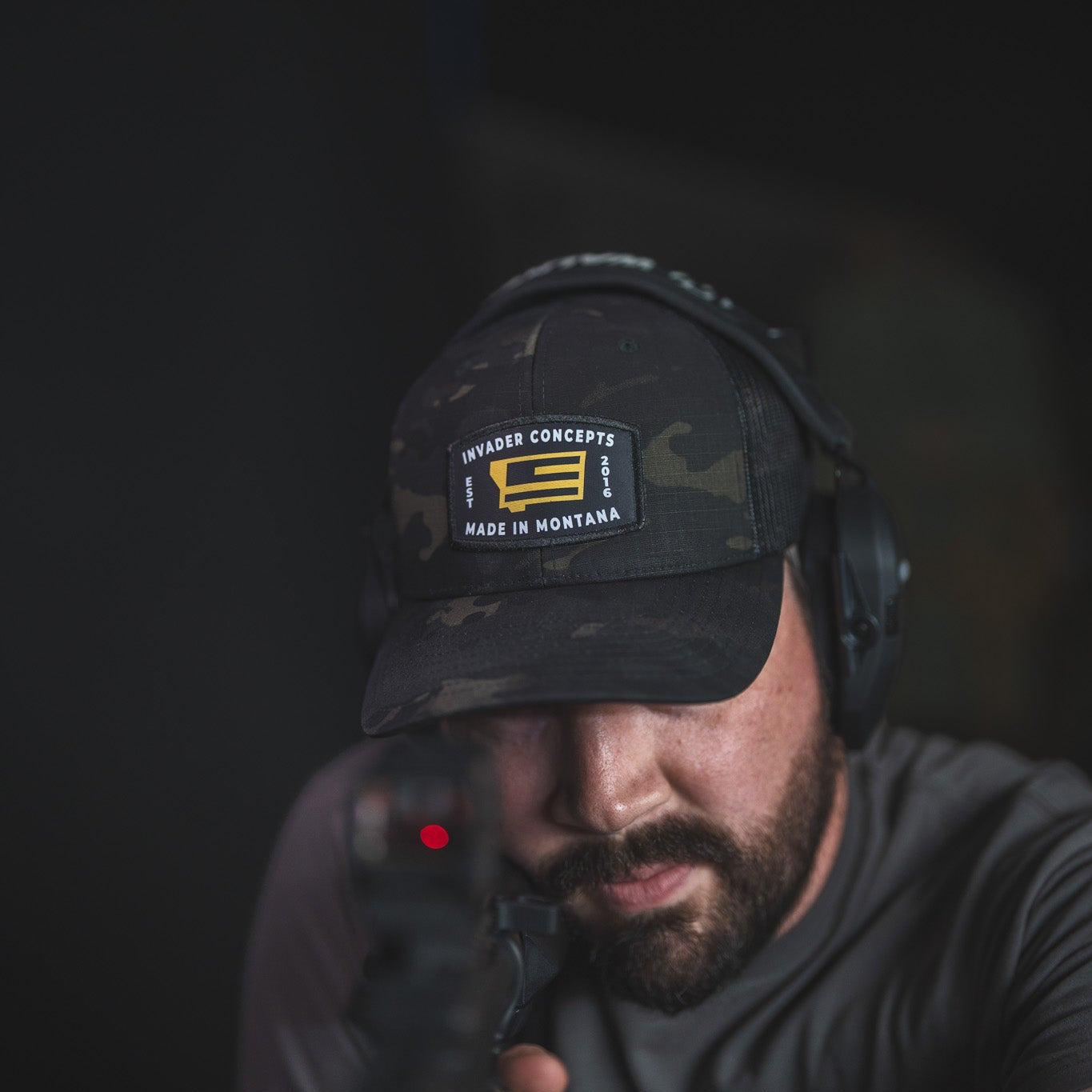 A man wearing a black camouflage baseball cap with a 'Hunter Conceal Made in Montana' patch, black over-ear headphones, and a dark gray t-shirt, aiming a handgun in a dimly lit setting.