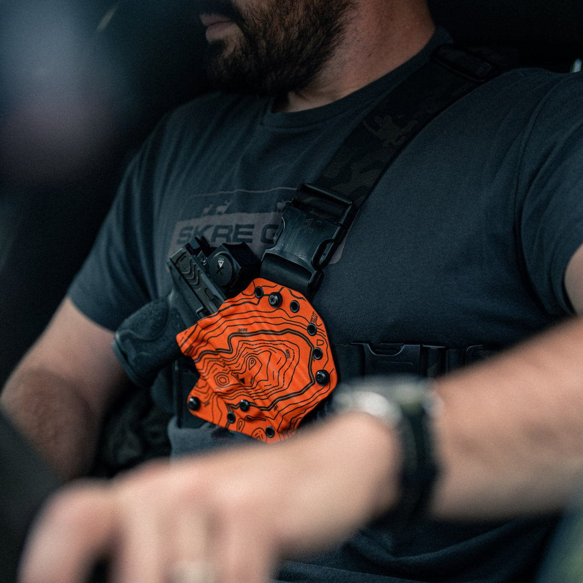 A close-up lifestyle shot of a man wearing a black tactical chest rig with an orange topographic map patch featuring a stylized face design.