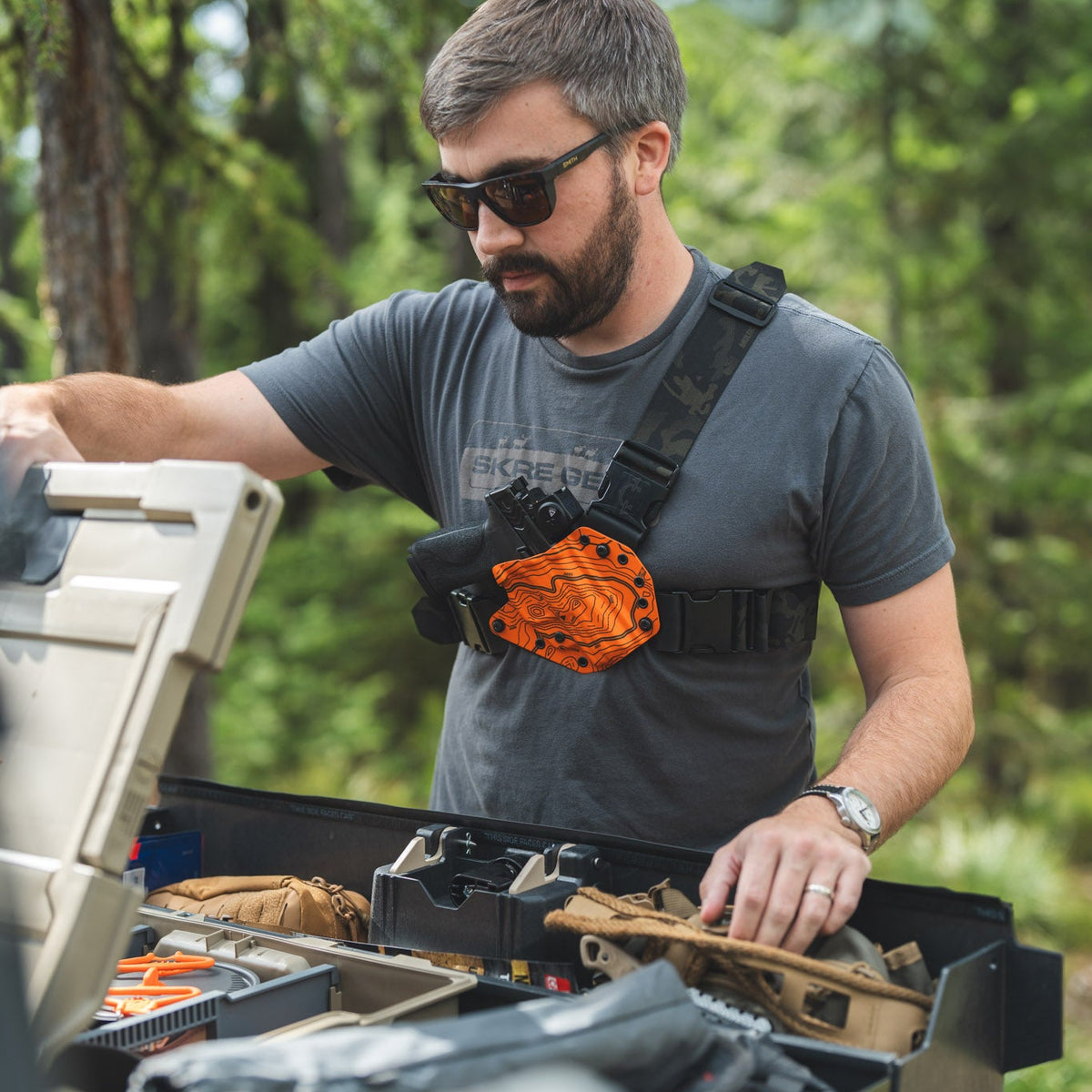A man wearing sunglasses and a gray t-shirt stands outdoors in a forest setting, organizing gear inside an open black storage case. He has an orange and black chest rig with a camera mounted on it.