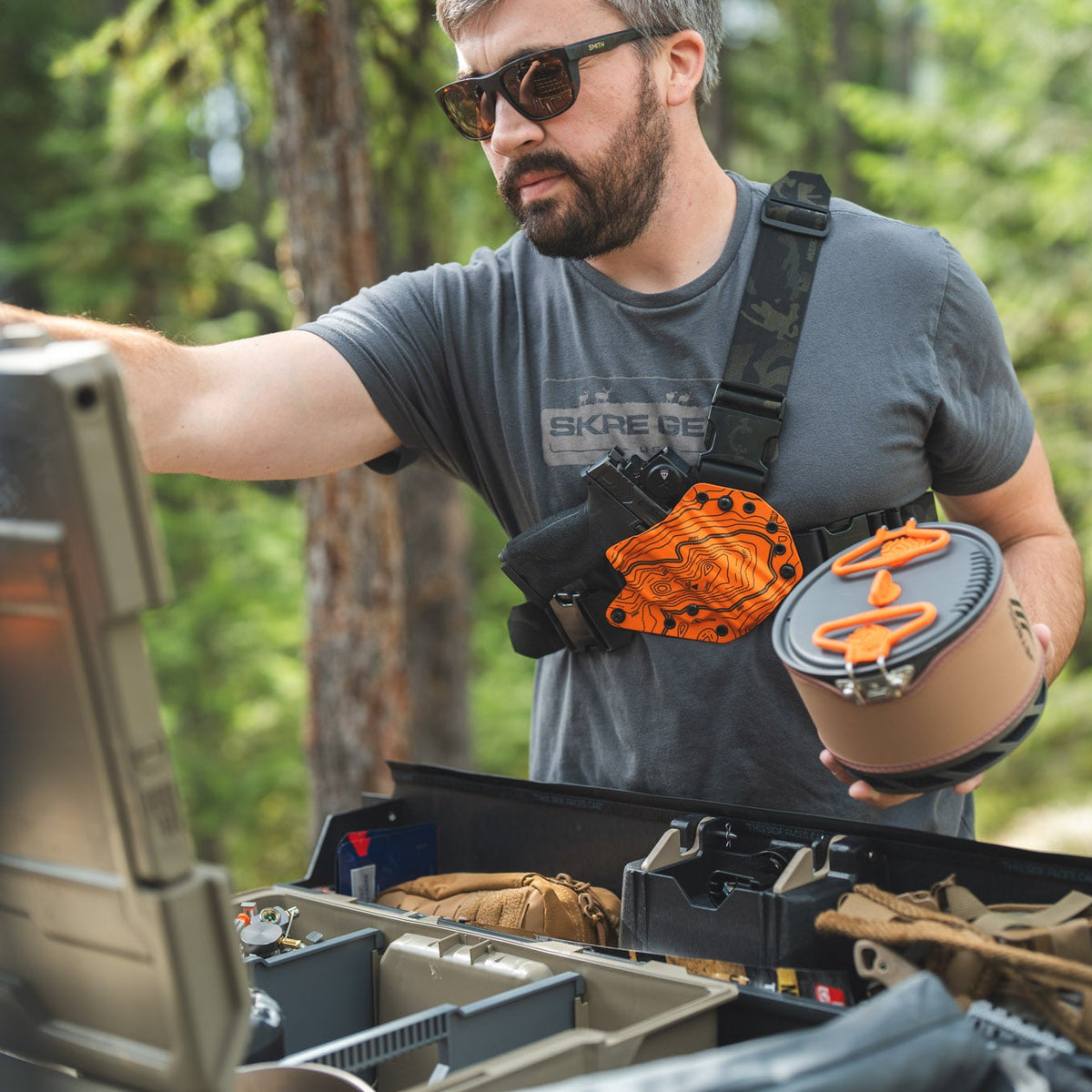 A bearded man wearing sunglasses and a gray t-shirt stands outdoors in a forest setting, holding a helmet with an orange logo while wearing a black chest rig with an orange pouch. He is reaching toward a black utility case in the foreground.