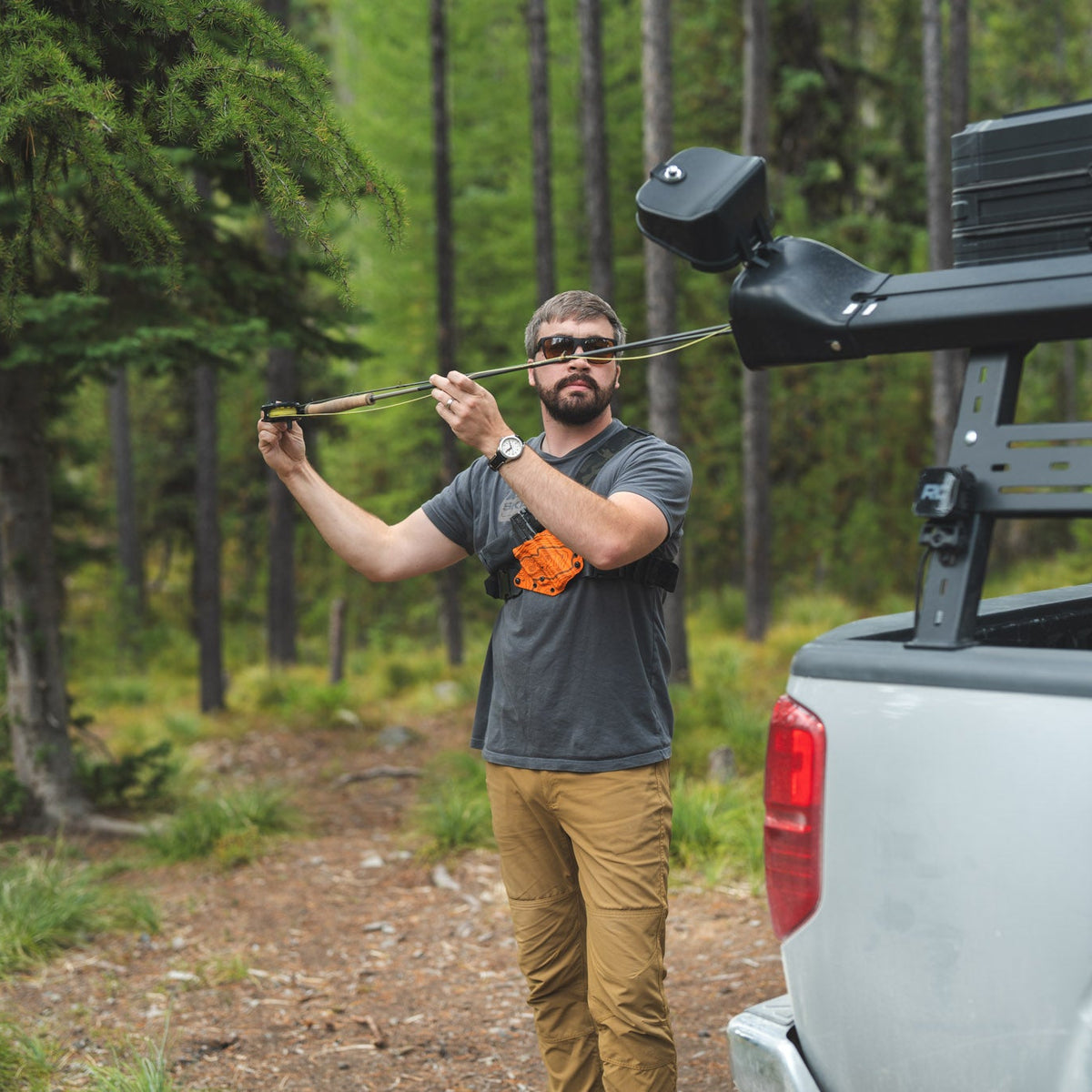 A man wearing a gray t-shirt and khaki pants stands on a forest dirt road next to a white pickup truck, holding a bow and arrow. The truck bed is equipped with a black roof rack and a mounted black storage box.