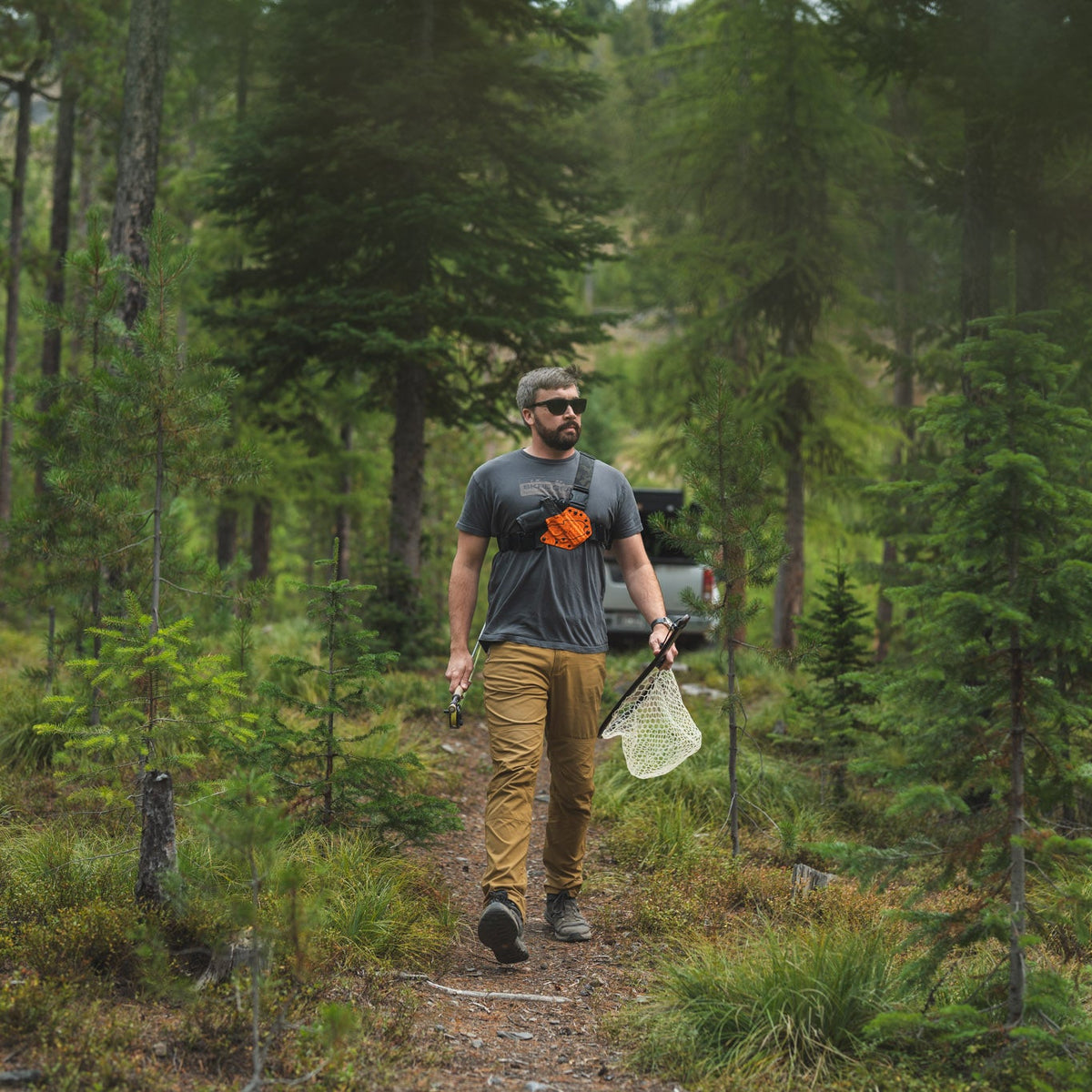 A man wearing a gray t-shirt with an orange logo and tan pants walks along a forest path carrying a white bag, with a vehicle visible in the background.