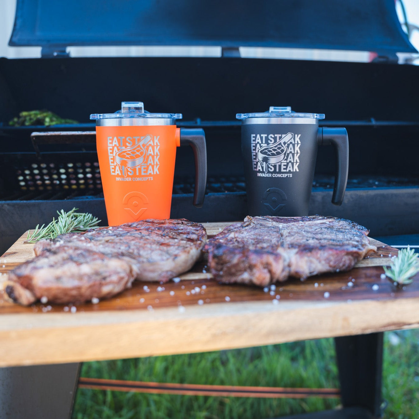 Two insulated tumblers, one orange and one black, both printed with the 'Eat Steak Steak Steak' logo, displayed on a wooden cutting board with two cooked steaks in front of a black smoker grill.