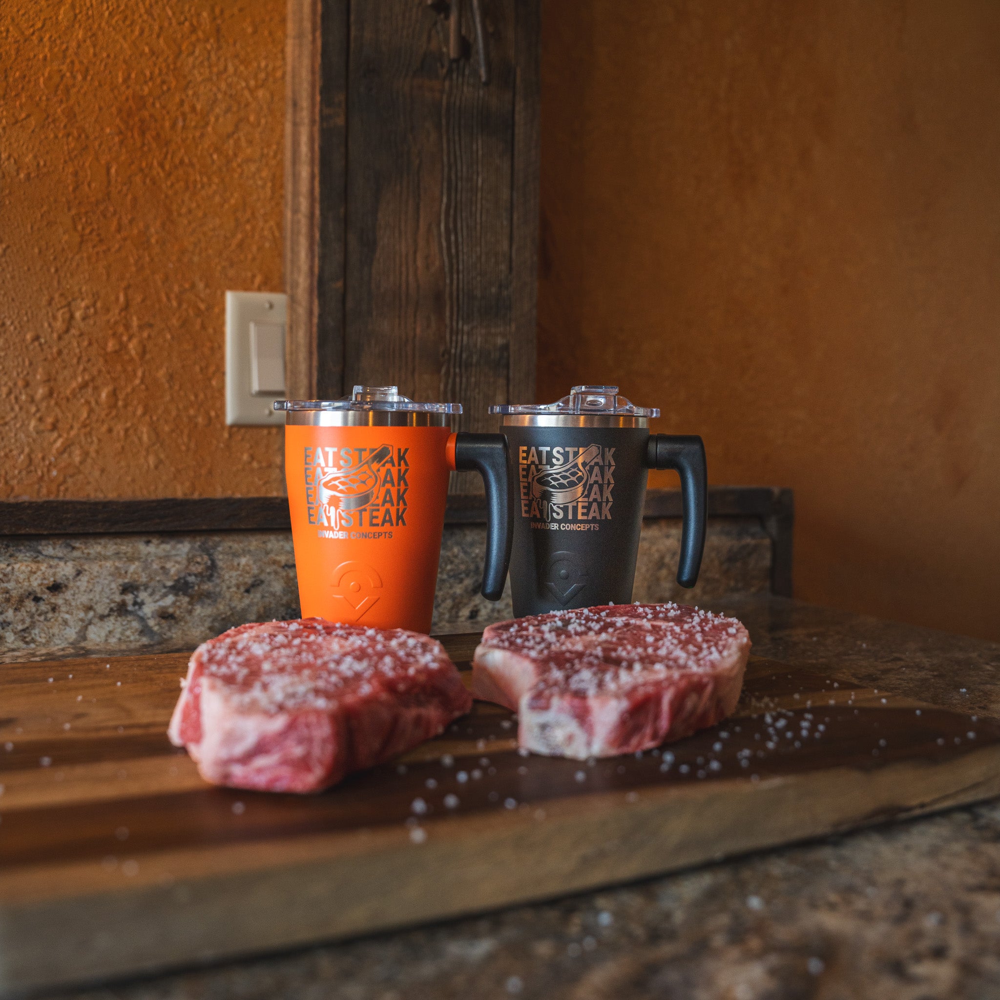 Two insulated travel mugs with lids and handles sit on a wooden cutting board in a rustic kitchen setting, with two raw steaks sprinkled with coarse salt in front of them. The mugs are one bright orange and one matte black, both printed with the Eat Steak Eat Steak logo.