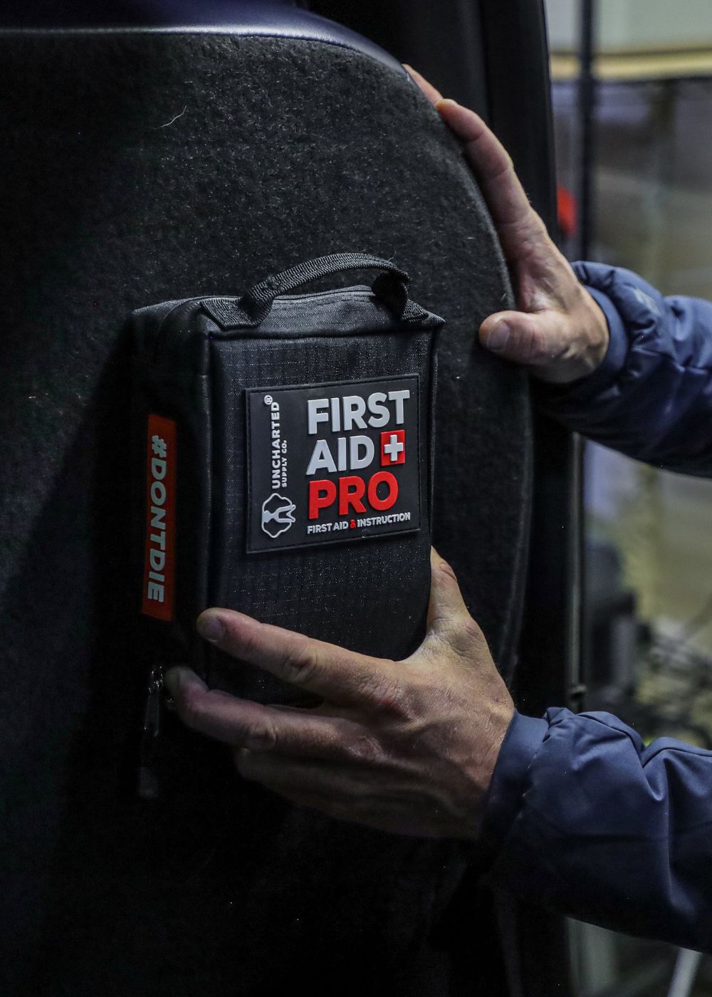 A black first aid kit pouch with a prominent 'FIRST AID PRO' label is being held by a person's hands against a dark textured surface.