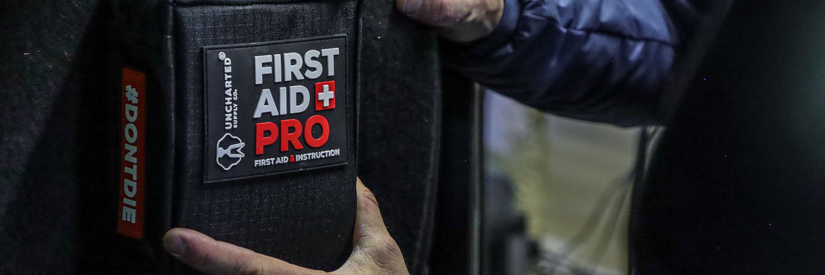 A close-up of a black first aid training patch on a dark fabric sleeve, with a person's hands holding it in place. The patch reads 'FIRST AID PRO' and 'FIRST AID / INSTRUCTION'.
